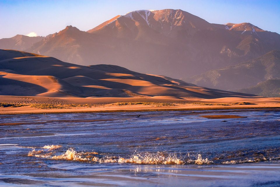 A small breaking wave crossing a shallow sheet of water in low golden light, dunes and a rosy mountain behind