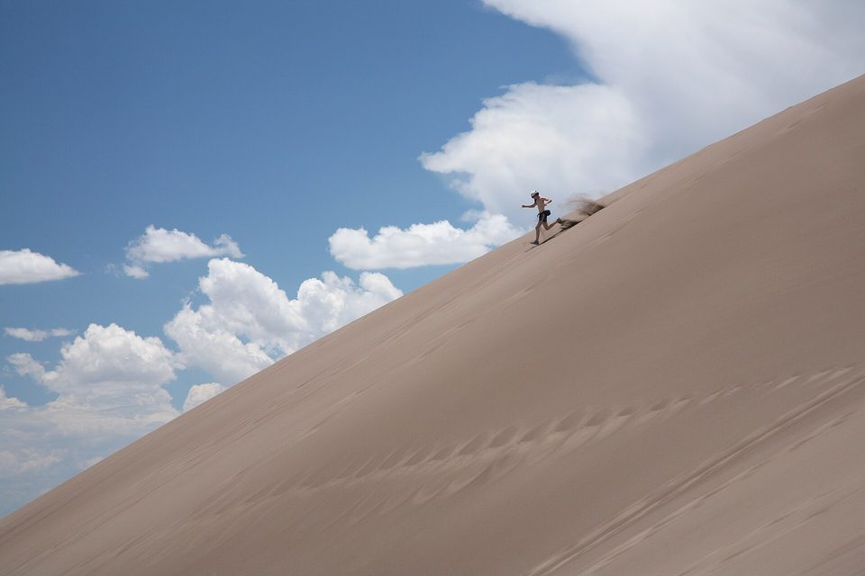 A lone figure running down a steep, smooth diagonal of sand under a blue sky with white clouds, sand spraying from their heels