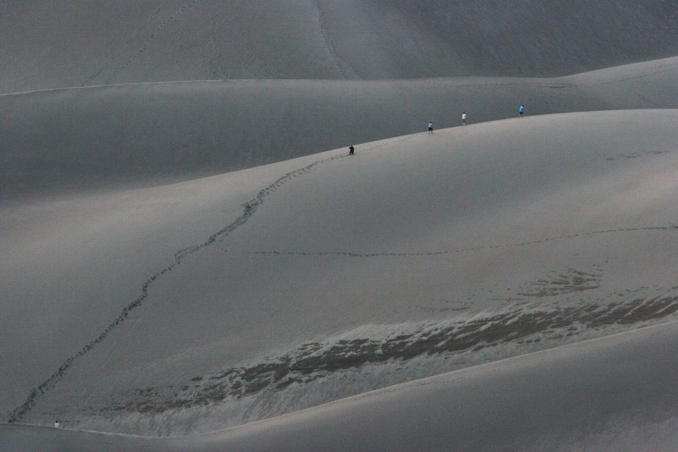 Four tiny walkers on the crest of a grey dune in flat evening light, a single line of footprints winding up the slope