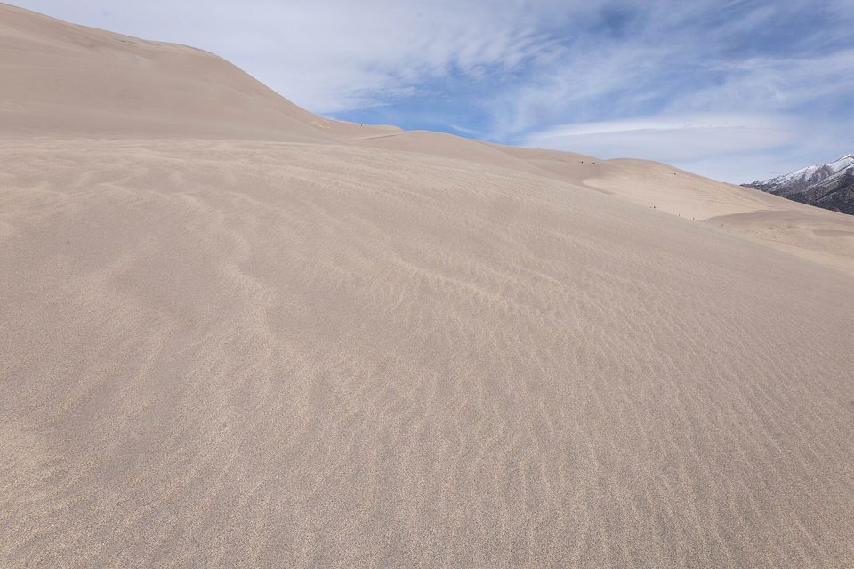 A broad pale sand slope covered in fine wind ripples, rising to a crest, snowy peaks just visible at right