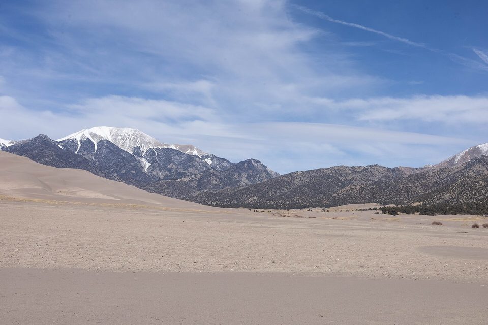 A flat pale plain of sand and scrub at the edge of the dunes, with snow-capped dark mountains behind under streaked cloud