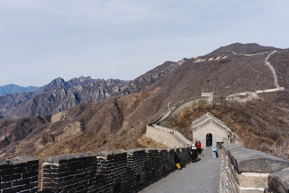A restored brick wall climbing a dry ridge to a gatehouse, with a few small visitors on the walkway and rugged peaks behind