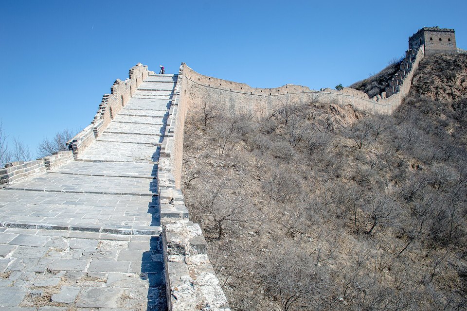A stone-paved wall walkway rising steeply between parapets toward a distant watchtower under a blue sky