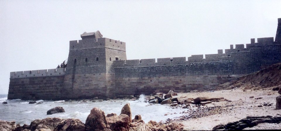 A tall grey brick tower and wall standing at the water line, with waves breaking over rocks and a pale sky