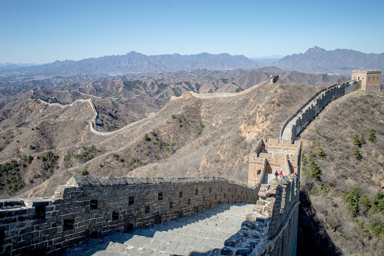 A restored stone wall with a watchtower climbing a bare ridge, more wall stretching over layered blue-grey mountains under a clear sky