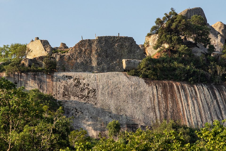 Walls of the Hill Complex built onto a streaked granite hill above the trees, in warm evening light under a clear sky
