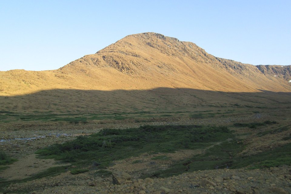 A bare, rust-orange mountain glowing in low evening sun above a shadowed valley of green shrubs and stones
