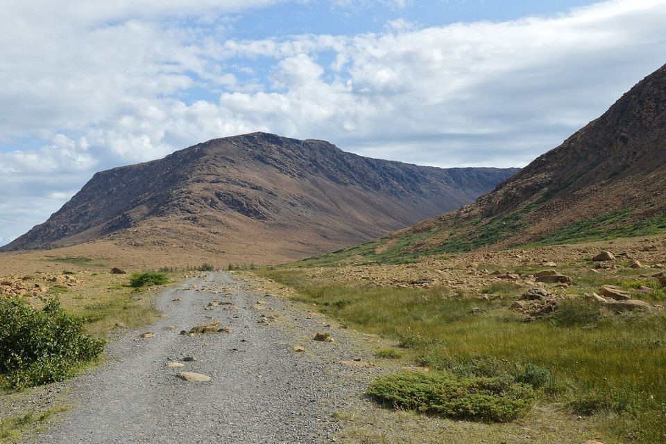 A gravel track running between low shrubs and scattered orange boulders toward a bare brown mountain under a cloudy sky