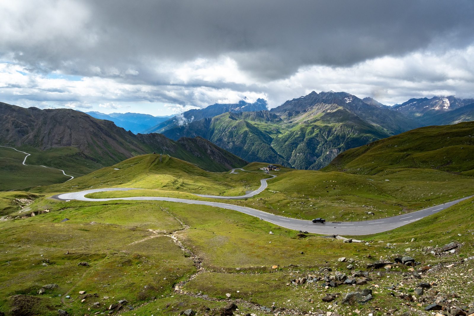 The Grossglockner road sweeping in long bends across high green pasture toward a mountain hut, jagged peaks catching sun under a dark storm sky