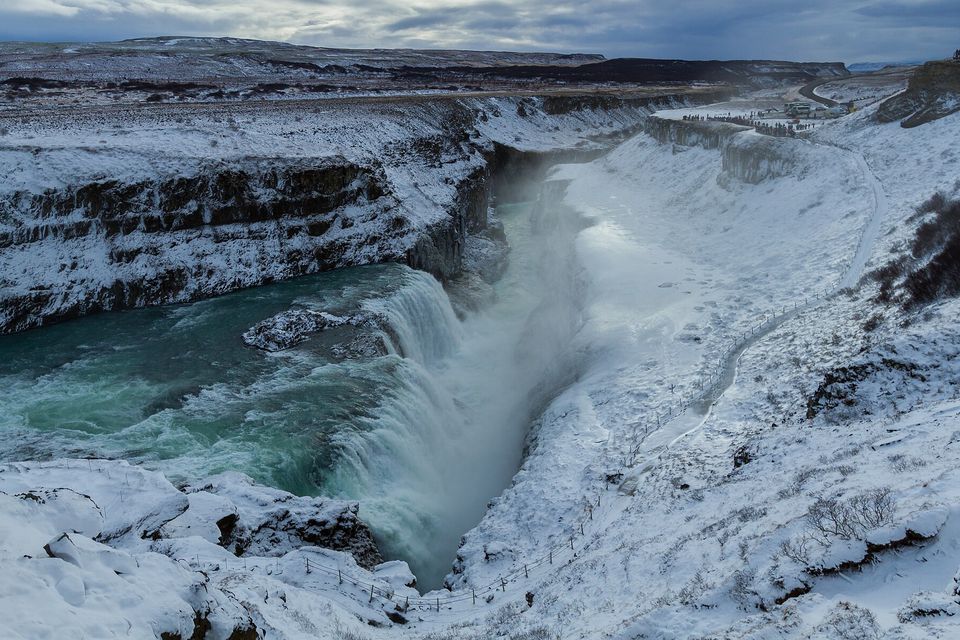 A wide snow-covered canyon with a green-grey river dropping over a fall at the left and spray rising from the gorge under a dark winter sky