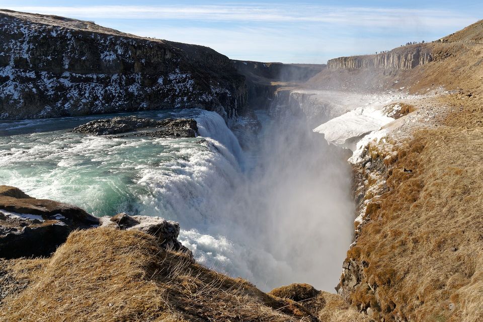 White water pouring over a rock lip into a narrow canyon filled with spray, snow-flecked dark cliffs at the left and a dry golden slope in the foreground