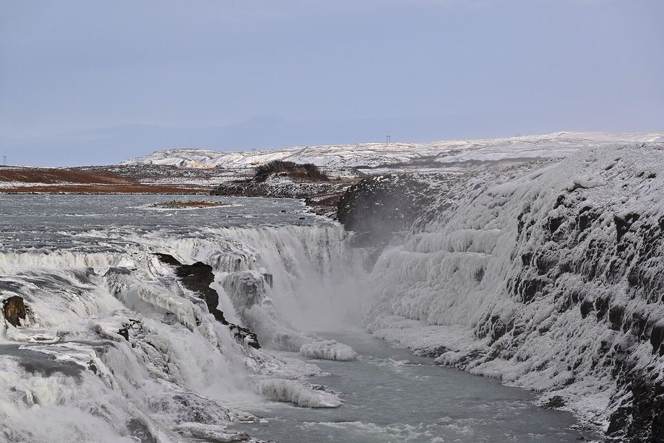 Grey-white water pouring down a stepped fall into a canyon, thick ice crusting the dark rock wall at the right under a pale grey-blue sky