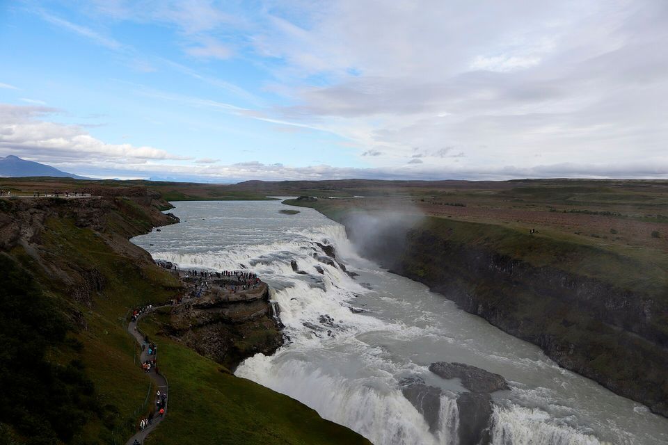 A wide river pouring over a stepped fall into a gorge below a grassy slope, a path winding down to a rocky ledge crowded with tiny visitors under a partly cloudy sky