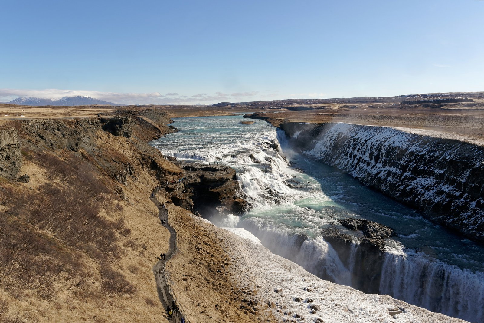 A wide glacial river turning and dropping in white cascades into a narrow canyon with snow-streaked walls, a path with a few tiny walkers on the left slope and a snow-capped mountain far off under a clear blue sky