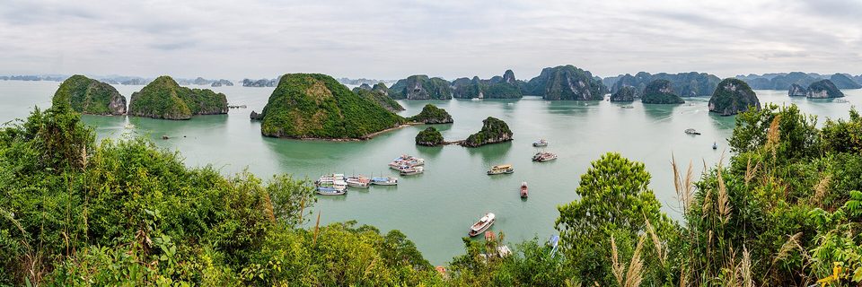 A wide view over green forested islets rising from calm jade water, tour boats moored below, layers of islands fading into haze