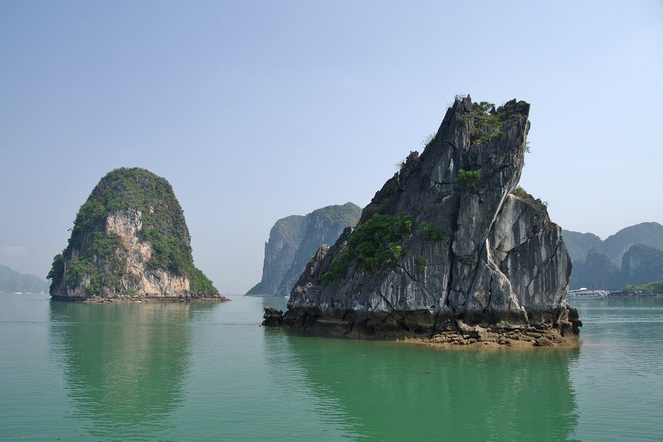 A jagged grey limestone tower whose base is cut back at the waterline, standing in turquoise water with another green-topped islet beside it