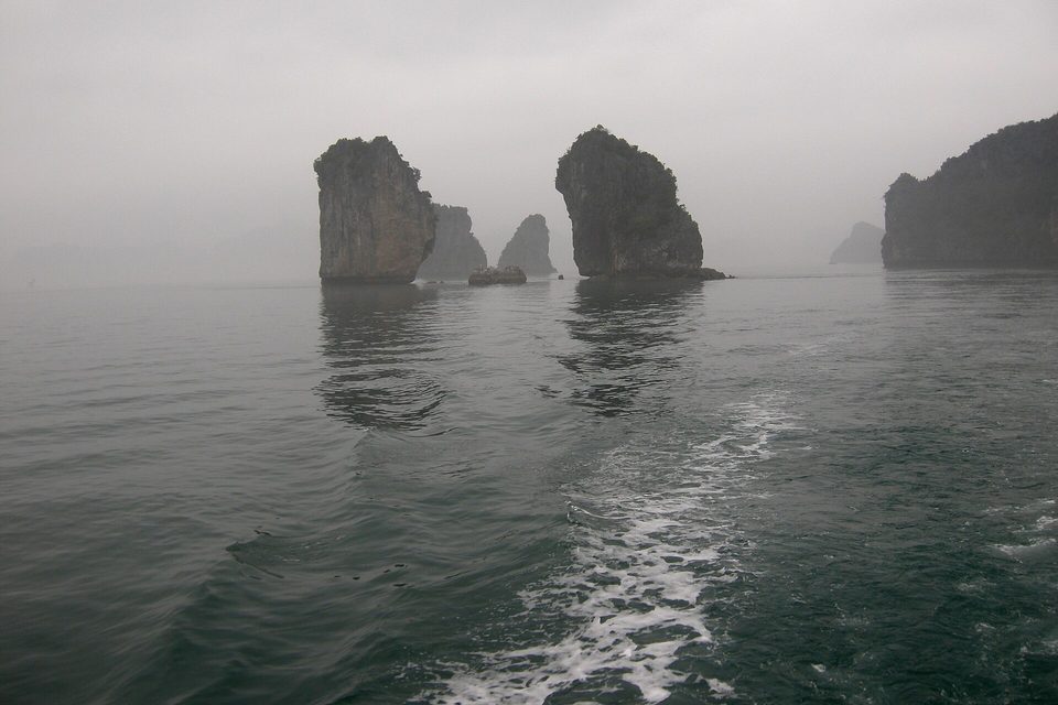 Several grey limestone towers standing in mist over dark water, a white wake running from the lower edge of the frame toward them
