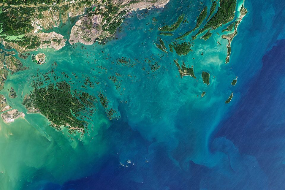 A satellite view of forested islands scattered through turquoise shallow water beside a built-up coast, deep blue sea at the right