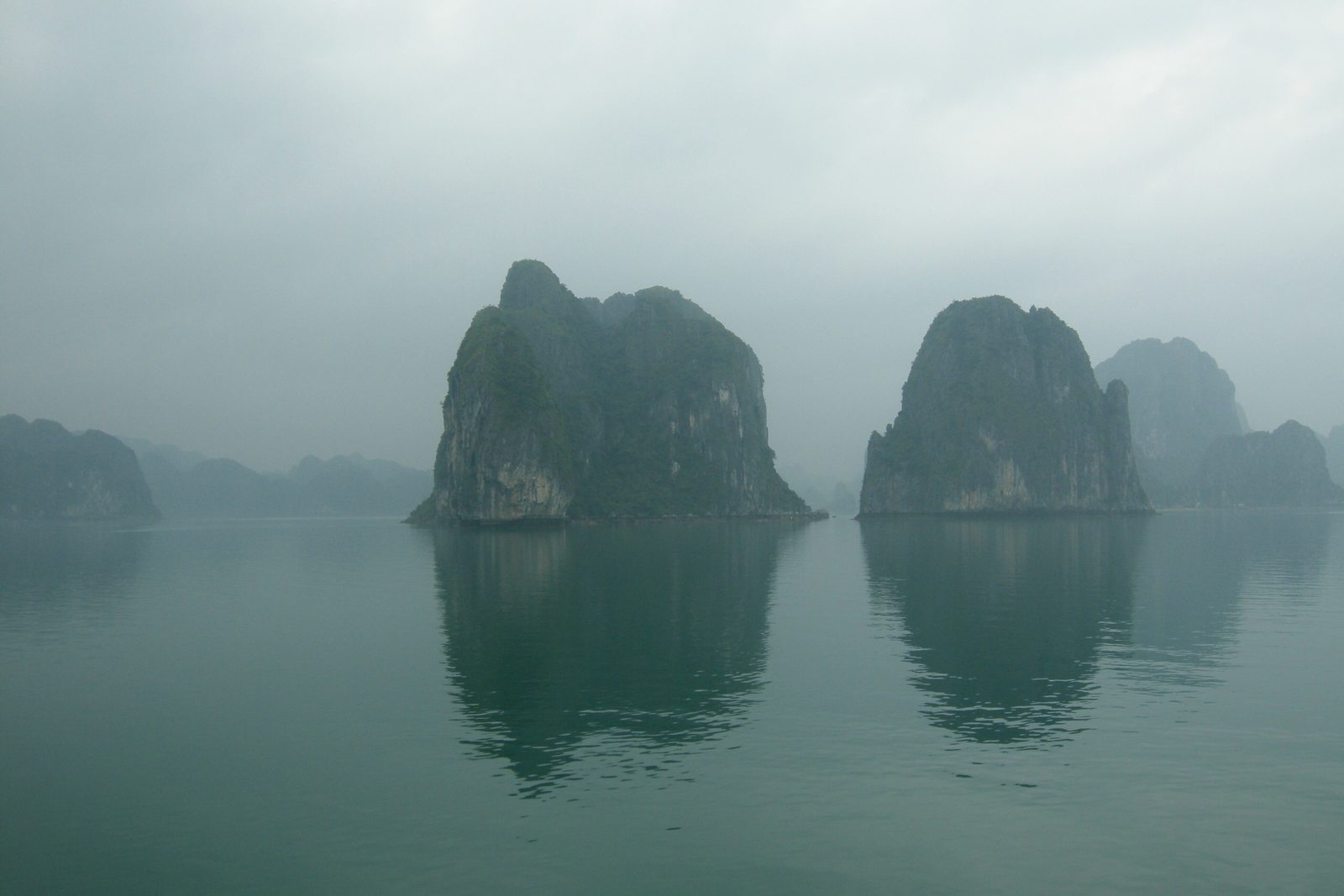Two steep green-grey limestone towers rising from flat, jade-coloured water under a soft grey sky, reflected below, more towers fading into mist