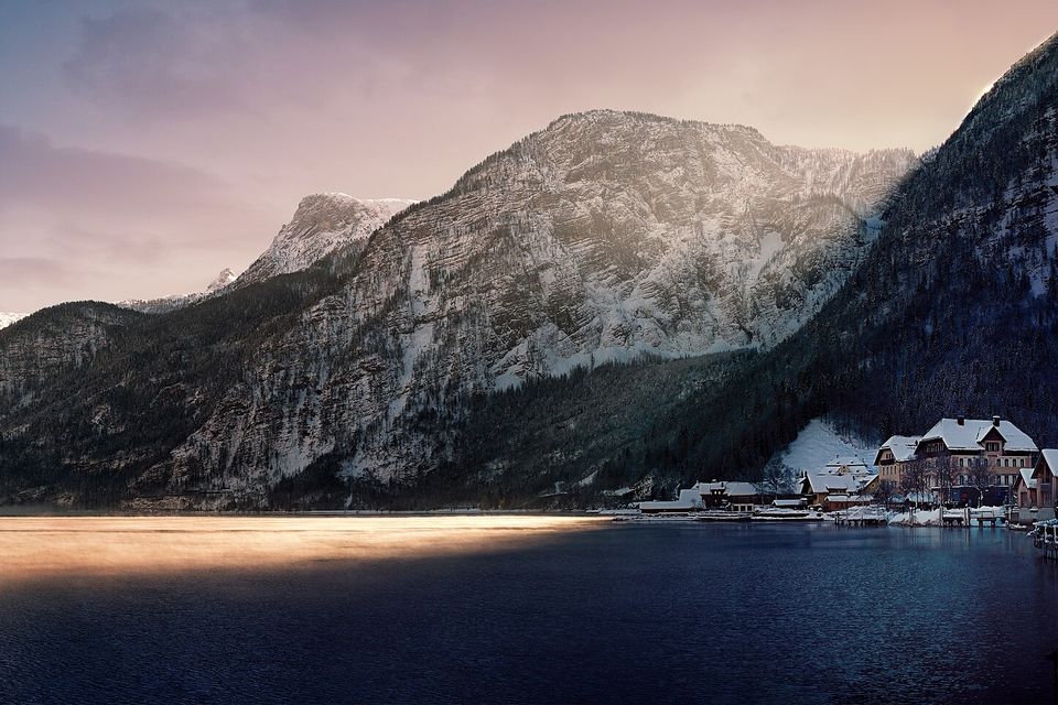Snow-dusted limestone mountains rise over a dark lake lit by a band of golden sunlight, the snow-covered village tucked under the cliffs at right
