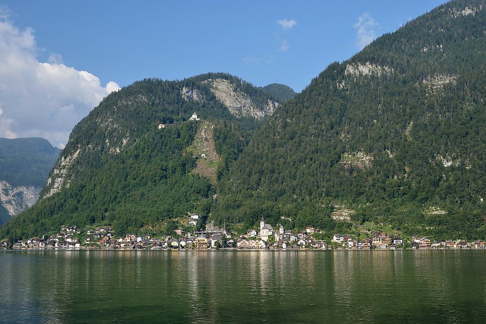 A thin line of houses and a church along a green lake shore, under steep forested mountains; a white tower and a cleared slope mark the high valley above the village