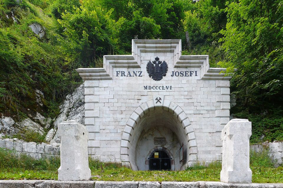 A pale stone portal carved with an inscription and a double-headed eagle above a gated arch, set into a wooded slope