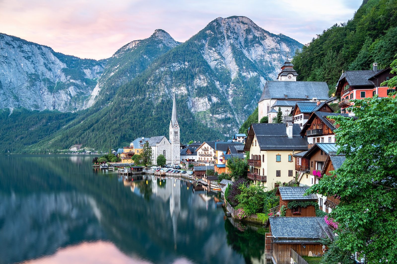 At dawn the slender spire of the Protestant church and the stacked houses on the shore are mirrored in a glass-still lake, pale limestone peaks under a pink sky