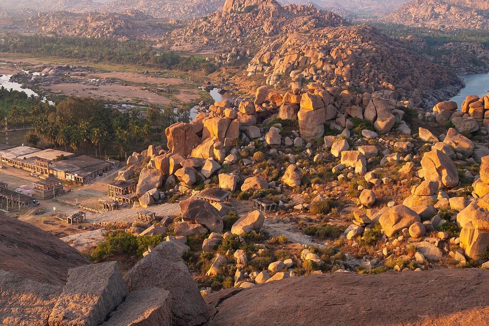 Golden-lit piles of granite boulders on a hillside above a river, a long colonnade and palm groves below and more boulder hills beyond