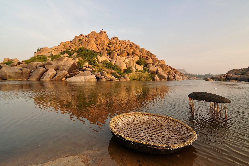 A round woven basket boat at the edge of calm river water, a second boat upturned on stilts, a boulder hill reflected in the river at dawn