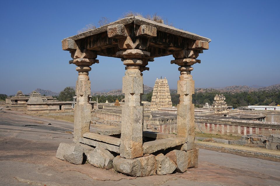 A four-pillared stone pavilion on bare rock, a tall temple gateway tower and temple compounds in the distance under a deep blue sky