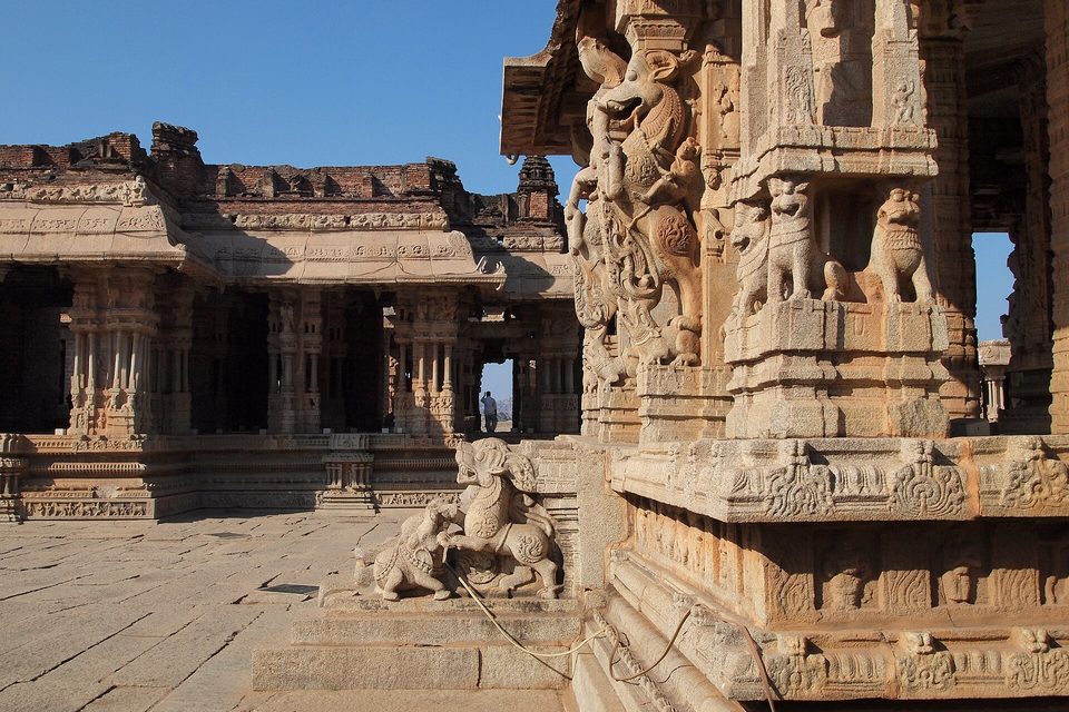 Carved granite pillars with rearing beast sculptures at the right, a pillared hall beyond and a stone rearing animal at a stairway, under blue sky