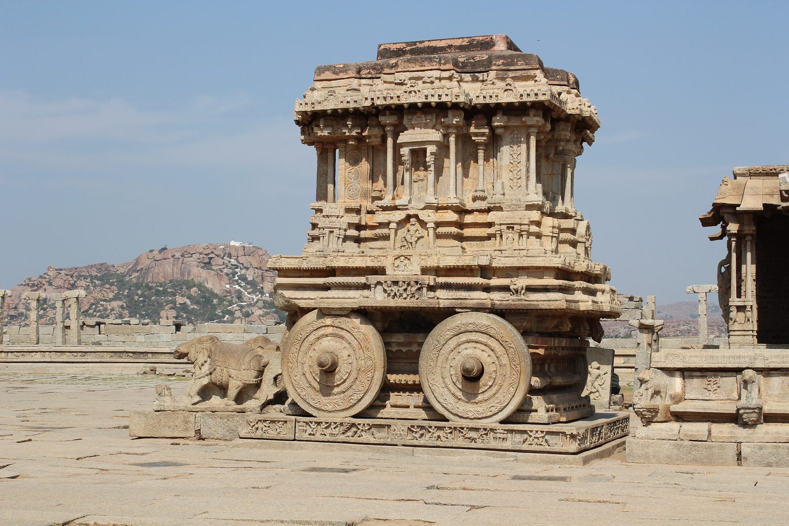 A carved stone chariot shrine on a stone plinth with two round stone wheels and an elephant carving at its front, a boulder hill and open pavilions behind, under a pale blue sky