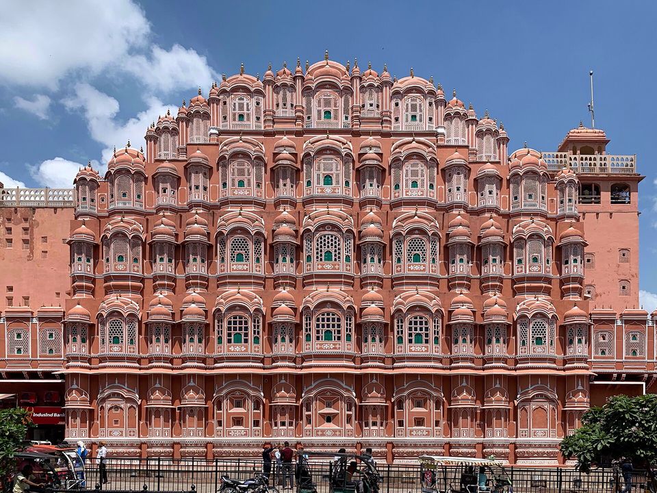The tiered pink facade of the Hawa Mahal in full under a blue sky with clouds, a railing and a few street figures and rickshaws at its foot