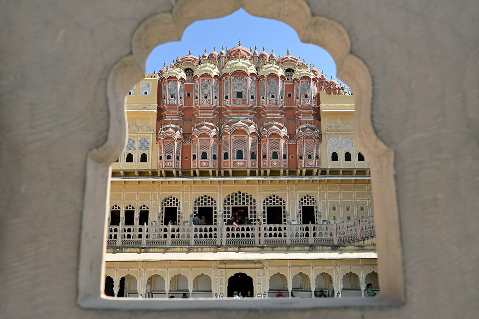 A scalloped stone window frame surrounding a view of the tiered pink and cream facade of the Hawa Mahal, with a colonnaded gallery below