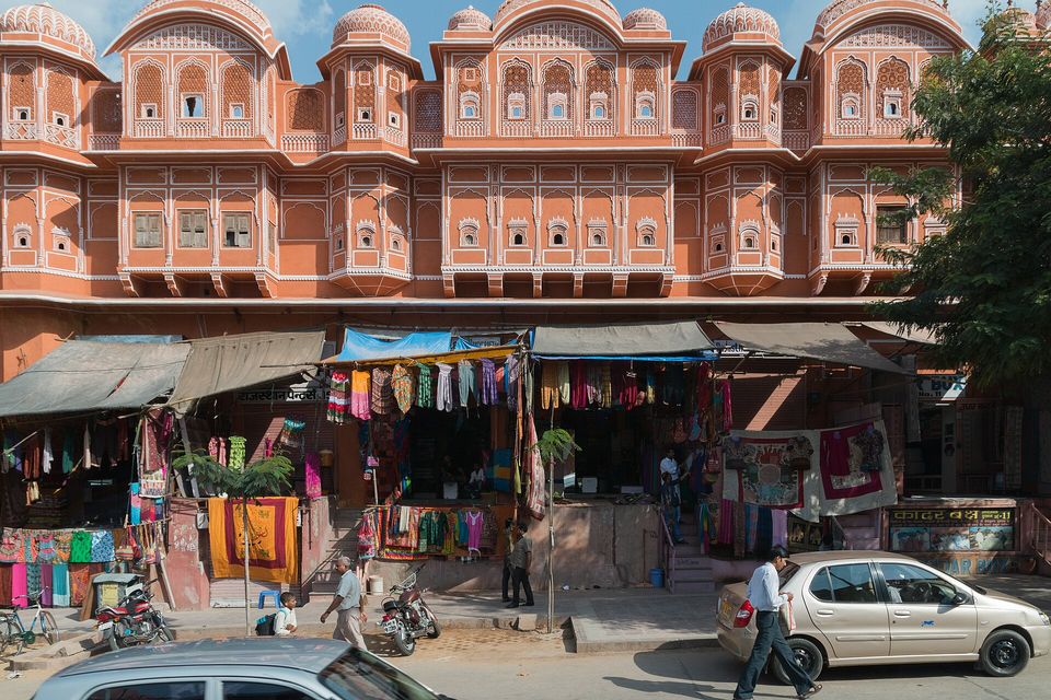 Shop stalls hung with bright cloth beneath the pink upper floors of an old-city building, a car and two walkers in the street