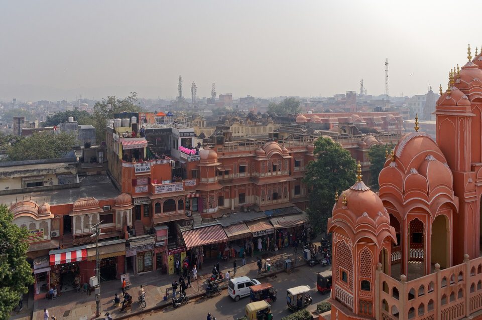 A street of pink and red shops and stalls seen from above, with motorbikes and auto-rickshaws, and the carved domed pavilions of the Hawa Mahal in the right foreground