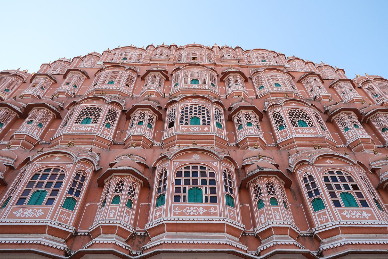 A five-storey pink sandstone facade covered in small latticed windows and scalloped balconies, seen from below against a clear blue sky