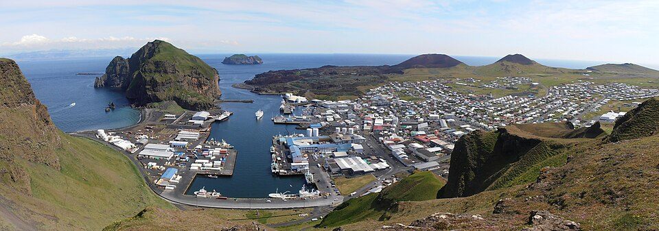 A deep natural harbour full of fishing boats lies below steep green cliffs, with a white town spreading up to two dark volcanic cones
