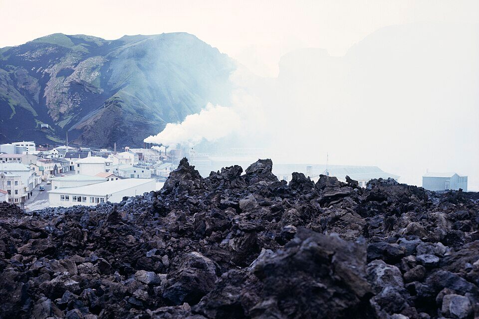 A field of jagged black lava fills the foreground while white steam drifts over the edge of a town of white buildings below a green mountain