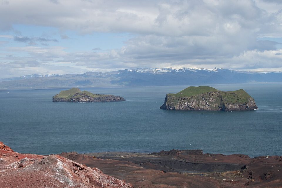 Two green-topped cliff islands sit in a grey-blue sea, with snowy mountains on the far shore and red cinder slopes in the foreground