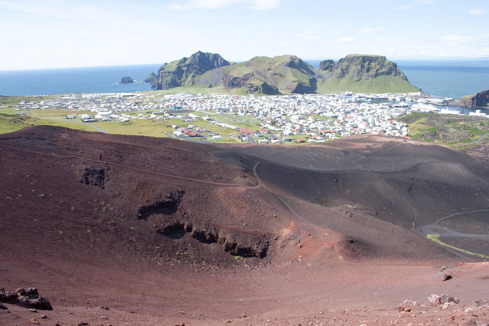 A red-brown cinder crater sweeps across the foreground, with a white harbour town, green cliffs and the sea beyond