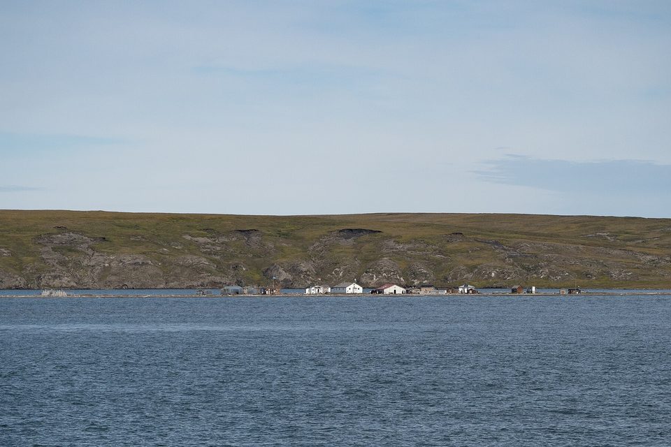 A thin line of white and red-roofed buildings along the shore, below low green hills scarred with bare, slumping patches, across blue water
