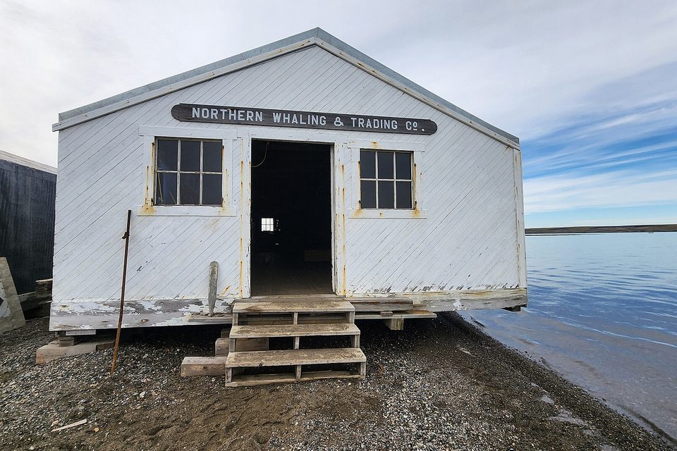 A weathered white clapboard building with a painted company name over an open doorway and wooden steps, on a gravel shore beside calm water