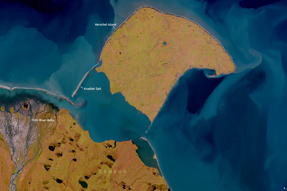 A satellite view of a rounded island of tan tundra in blue-green sea, with pale sediment plumes and a long spit to the west