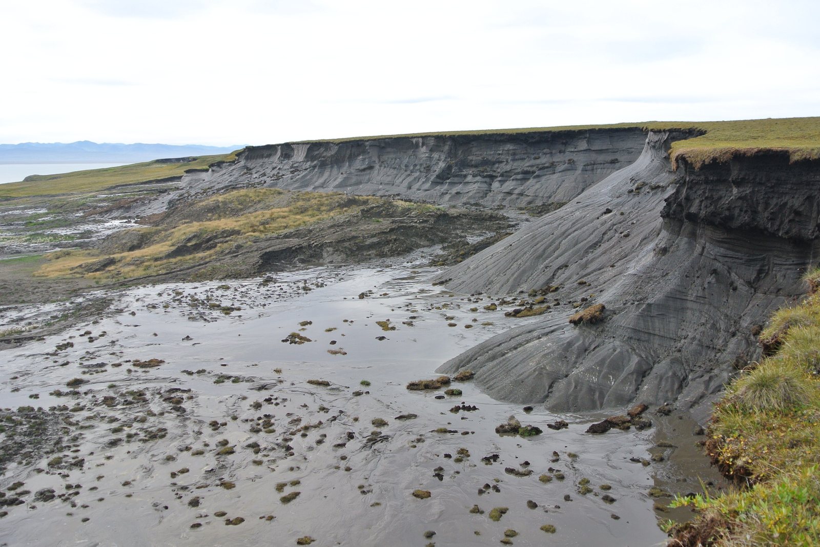 A grey headwall of ice-rich mud slumps from a lip of green tundra into a wet mudflow strewn with turf blocks, with the sea and distant mountains far left