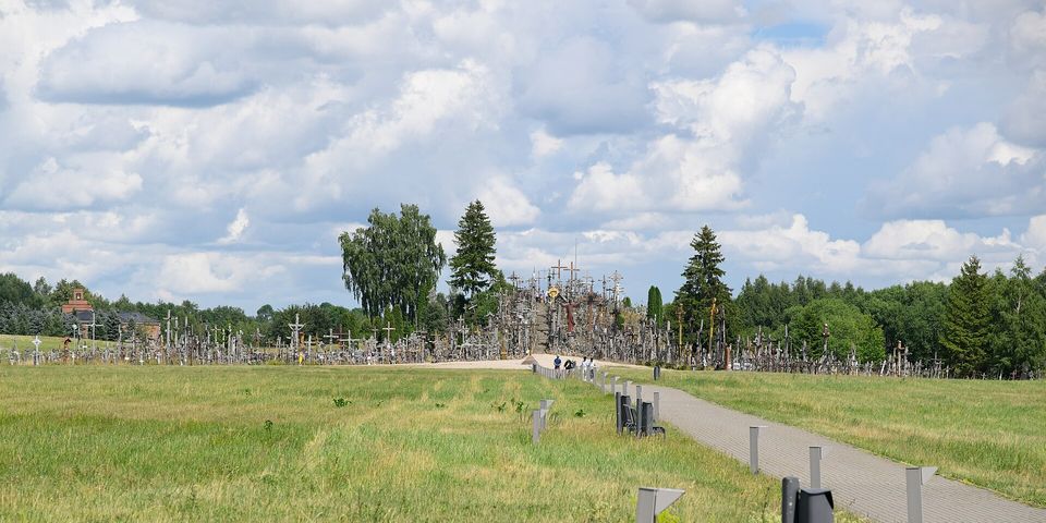 A low hill bristling with thousands of crosses rises beyond a long meadow and a paved path lined with benches, under a summer sky of white cloud