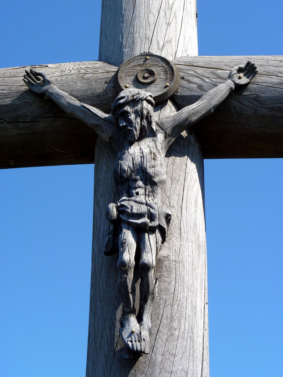 A silver-grey, weather-cracked wooden figure of Christ carved on a massive wooden cross against a deep-blue sky