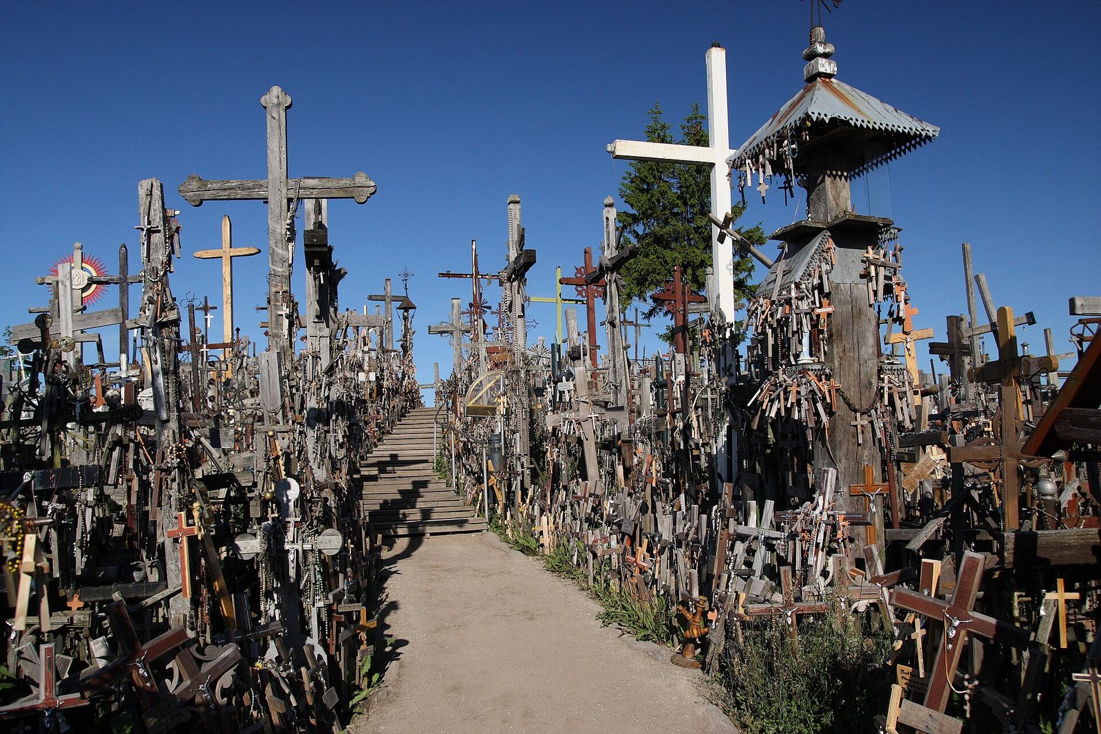 A sandy path runs up between walls of crosses, large and small, wooden and iron, hung with rosaries, to wooden steps climbing the hill, a white cross and a carved roof-pole shrine at right against a clear blue sky