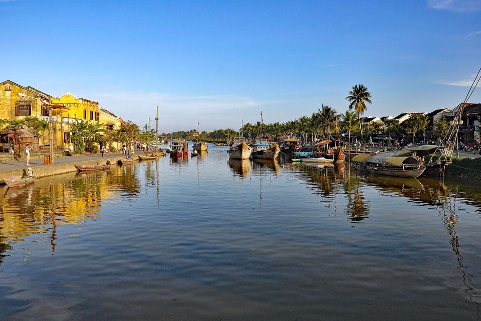 A calm river in warm light with wooden boats moored along both banks, yellow houses and palm trees on the quay and a blue sky above