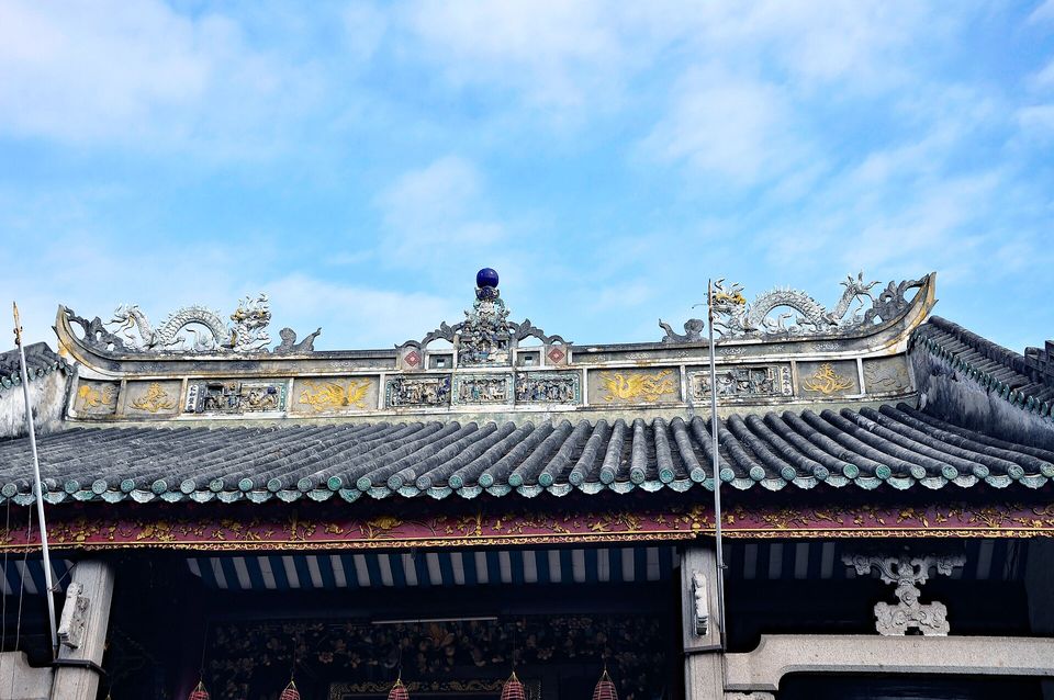 A tiled roof ridge decorated with two ceramic dragons and carved panels, a flagpole rising beside it, against a blue sky